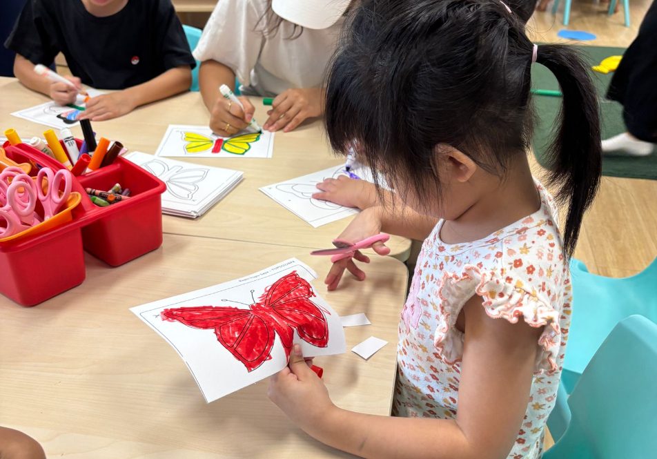 Child cuts out a red butterfly drawing at a table while others color butterfly sheets; markers and scissors nearby.